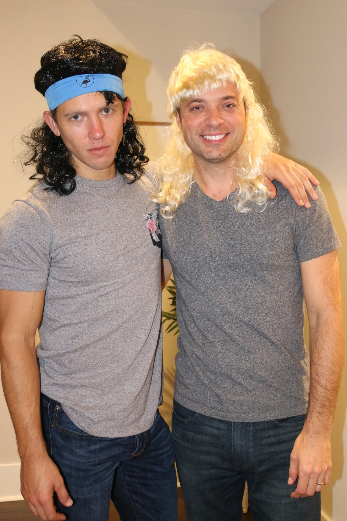 Two friends posing together, one in a black-curly mullet wig and blue headband, one in a blonde mullet wig