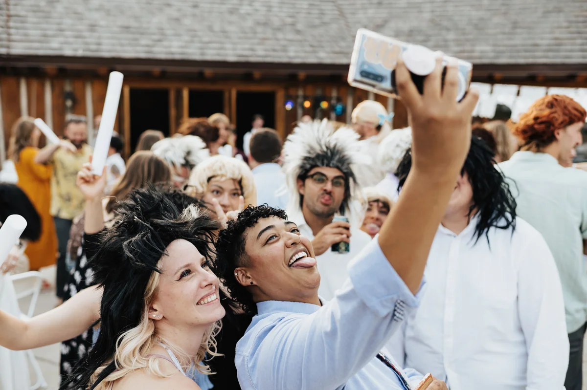 Two friends taking a selfie, tongues out, a row of mulleted guests behind them.